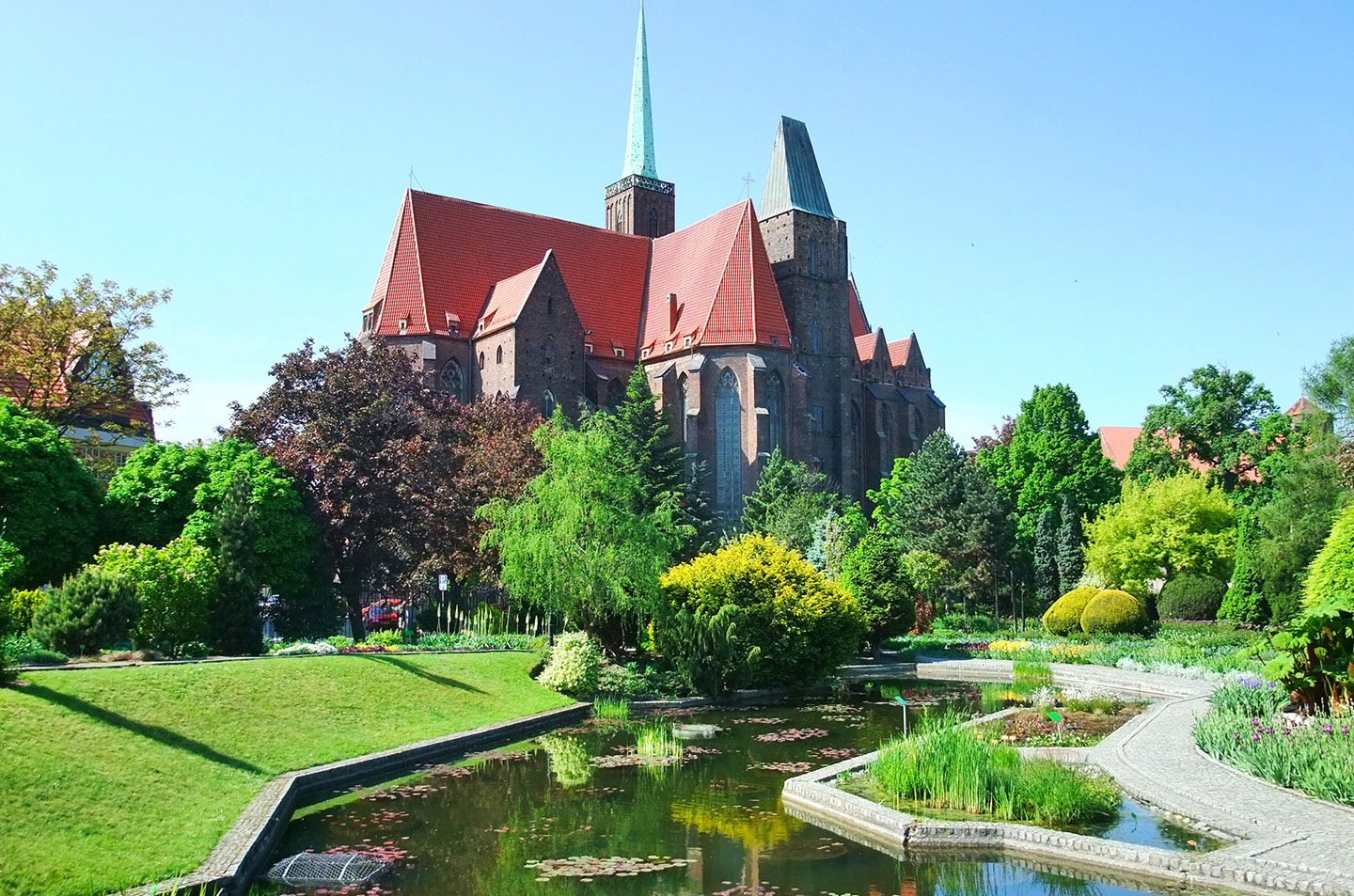 Manicured botanical garden with stone paths, flowering beds, and a reflective pond surrounded by mature trees.